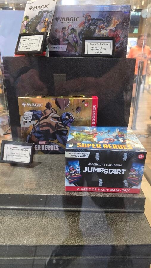 Stack of Magic The Gathering booster boxes including Jumpstart and Marvel Super Heroes on a display shelf in a store window