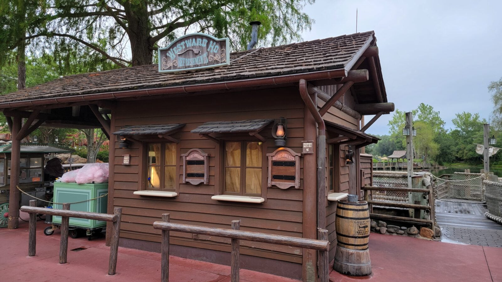 Brown wooden refreshment stand with signs reading Refreshments in a rustic park setting by a body of water and trees behind it