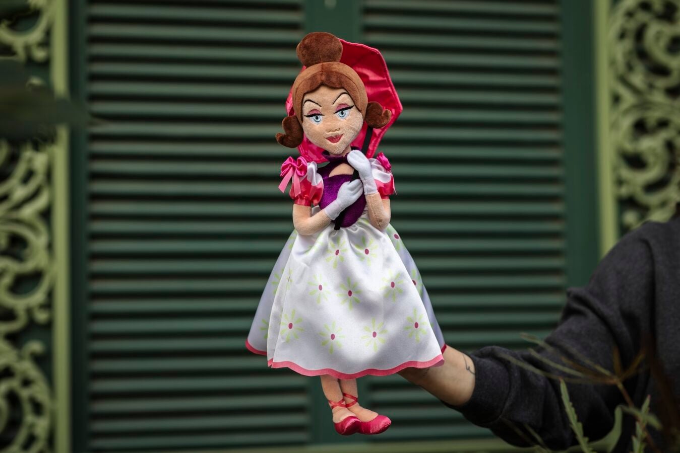 Hand held plush princess doll with brown hair bun pink cape and a white floral dress against green shutters