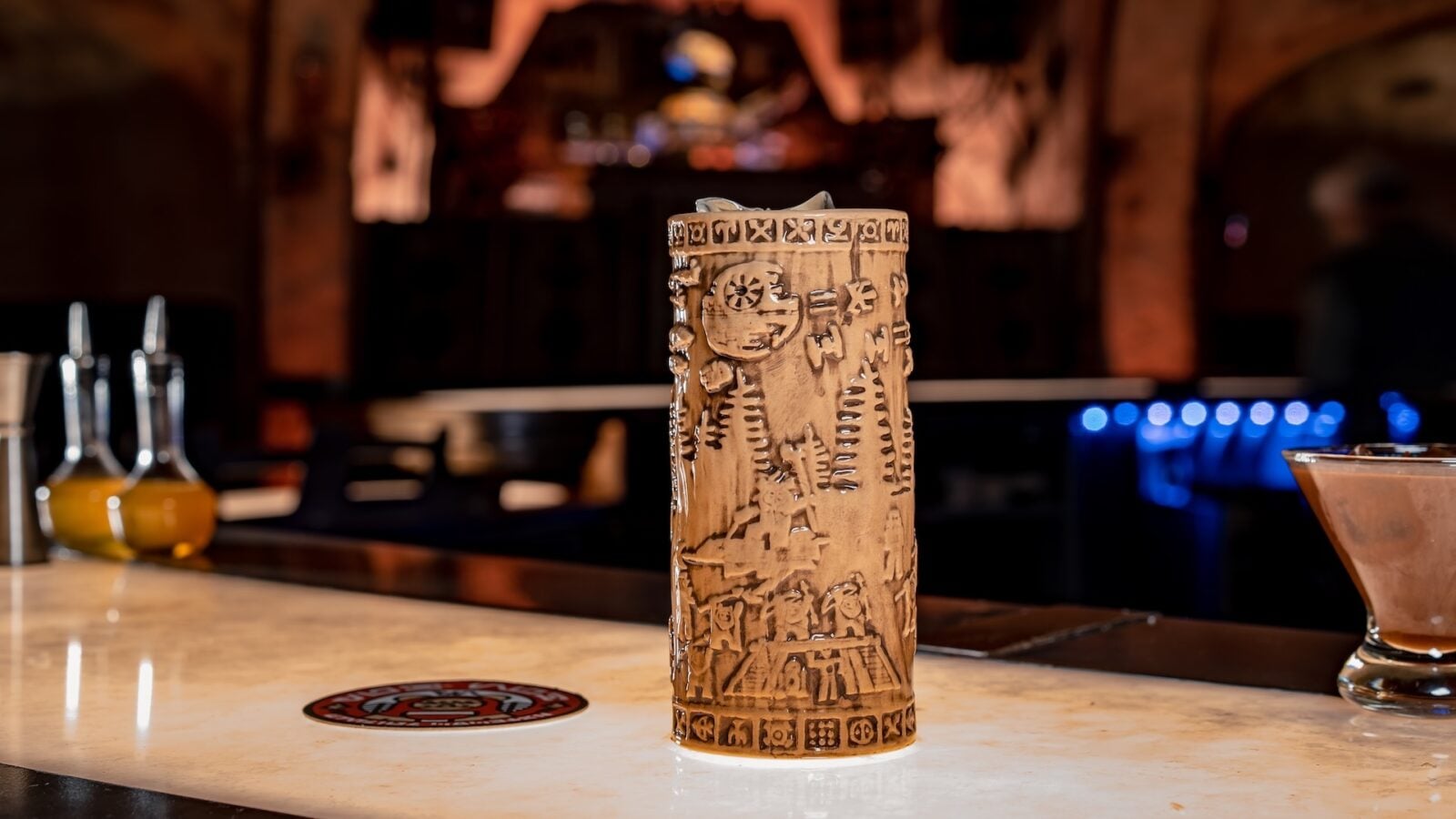 Tall carved wooden tiki mug with tribal patterns sits on a marble bar counter dimly lit bar in the background