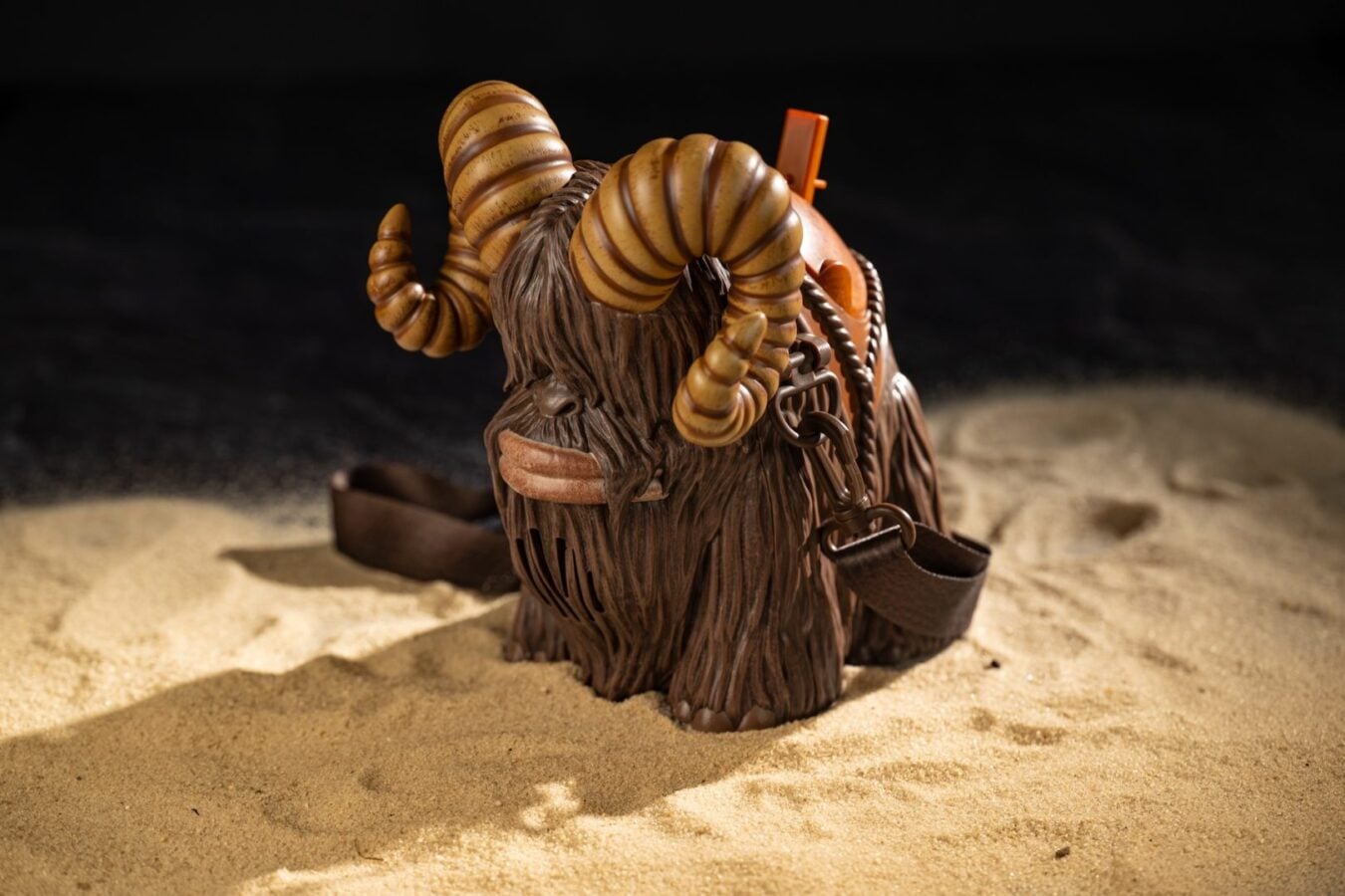 Brown shaggy figurine with large striped ram like horns wearing leather straps set on sandy surface against a dark background