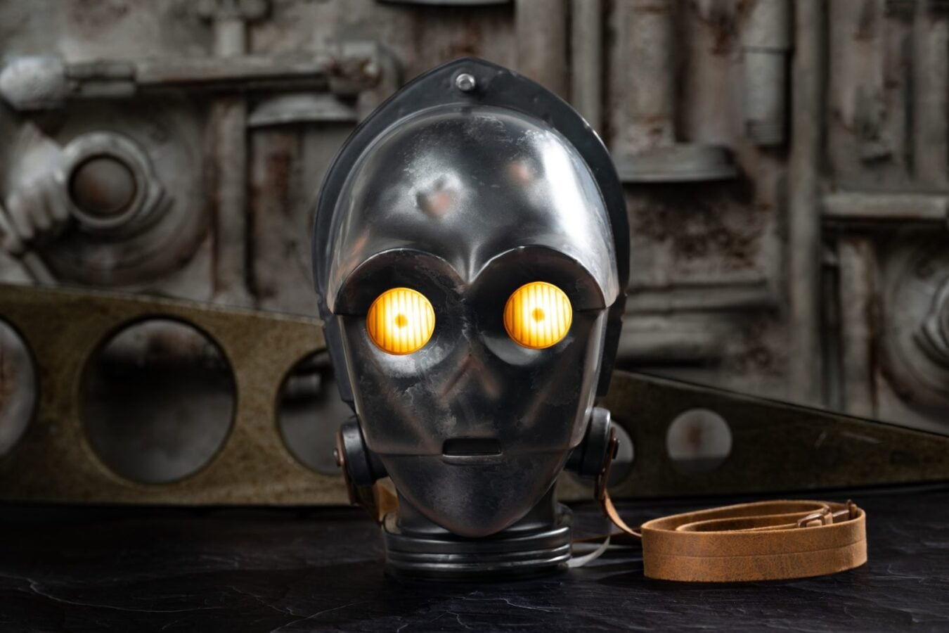Metal robotic head wearing a gas mask with glowing amber eyes in a dusty industrial workshop backdrop leather strap coiled nearby
