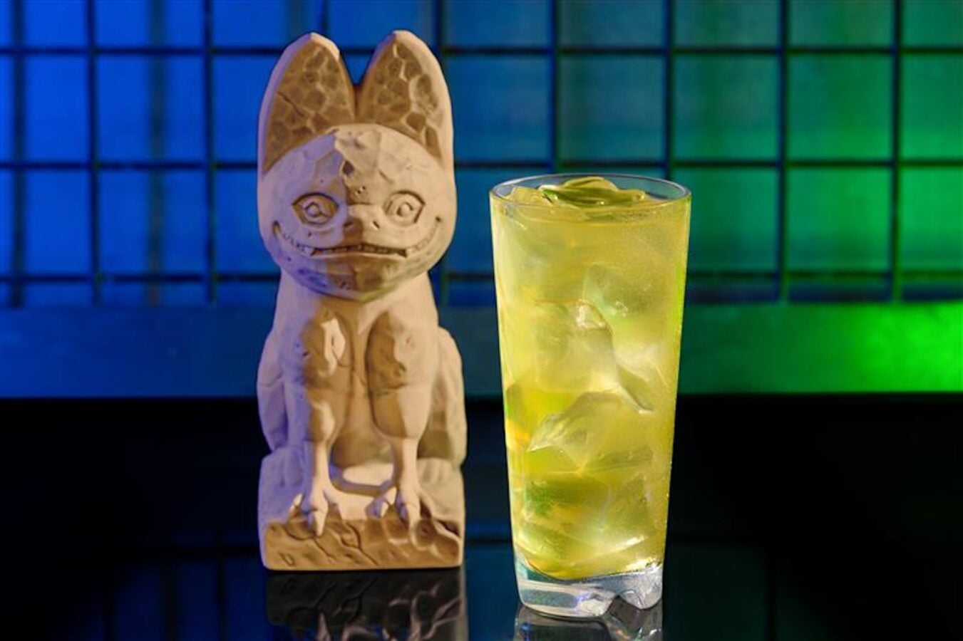 Stone dog figurine beside a tall glass of yellow iced drink on a reflective surface with a blue green tiled background