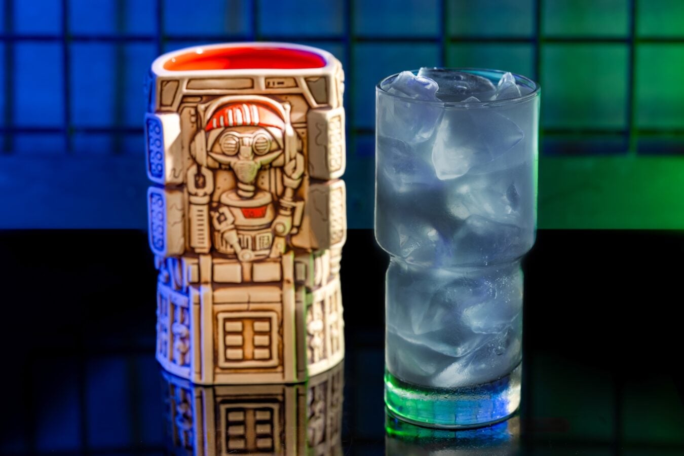 Ceramic mug with a robot inspired design beside a glass of ice water on a glossy surface with a blue green background