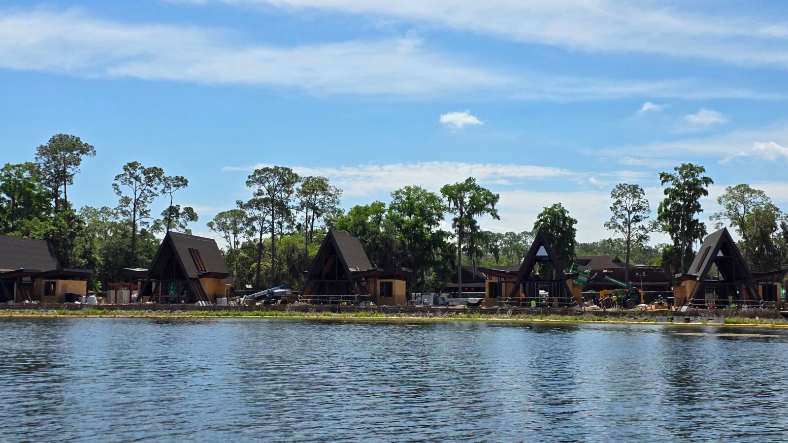 Lakeside construction site with dark triangular A frame buildings along the water and tall trees in the background