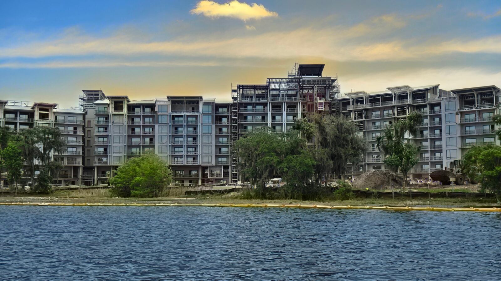 Modern apartment complex under construction along a river scaffolding visible and trees in the foreground