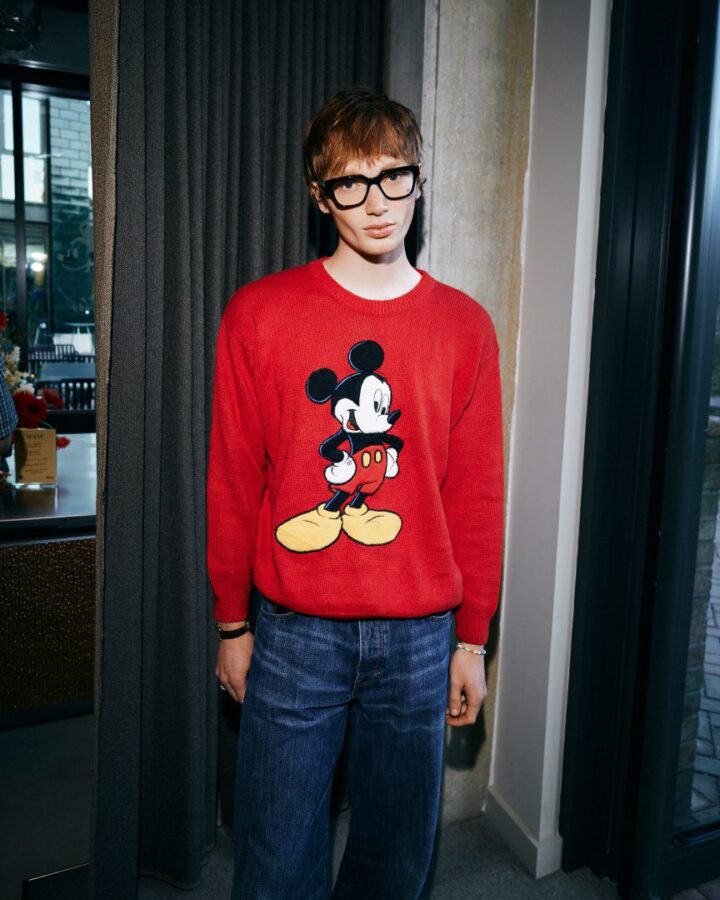 Person wearing a red Mickey Mouse sweater and blue jeans with black framed glasses standing indoors by a doorway