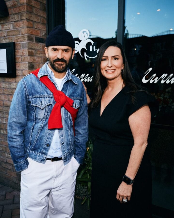 Man in a denim jacket with a red sweater tied over his shoulders wearing a beanie standing next to a smiling woman in a black dress outside a brick storefront