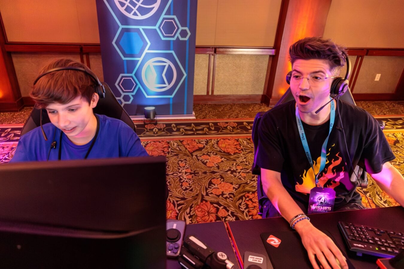 Two gamers sit at a competition station one in a blue shirt on the left and the other in a black shirt on the right wearing headsets with a blue geometric banner in the background
