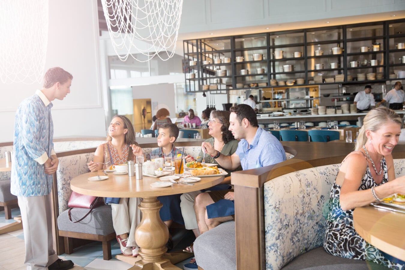 A waiter stands beside a booth chatting with a family enjoying breakfast or brunch at a bright modern restaurant