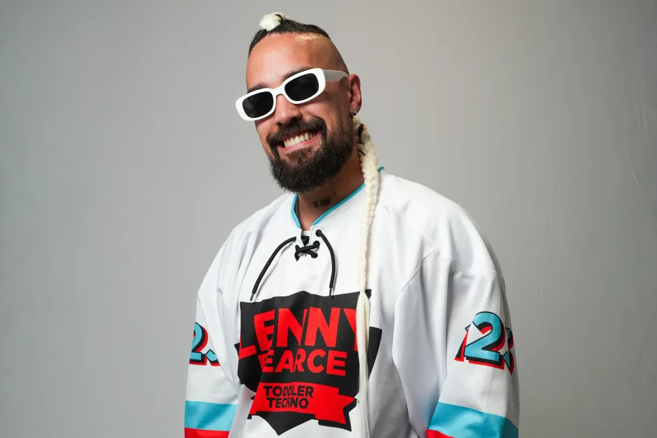 Smiling man with white sunglasses and a long braided ponytail wearing a white sports jersey with bold red lettering posing for a photo
