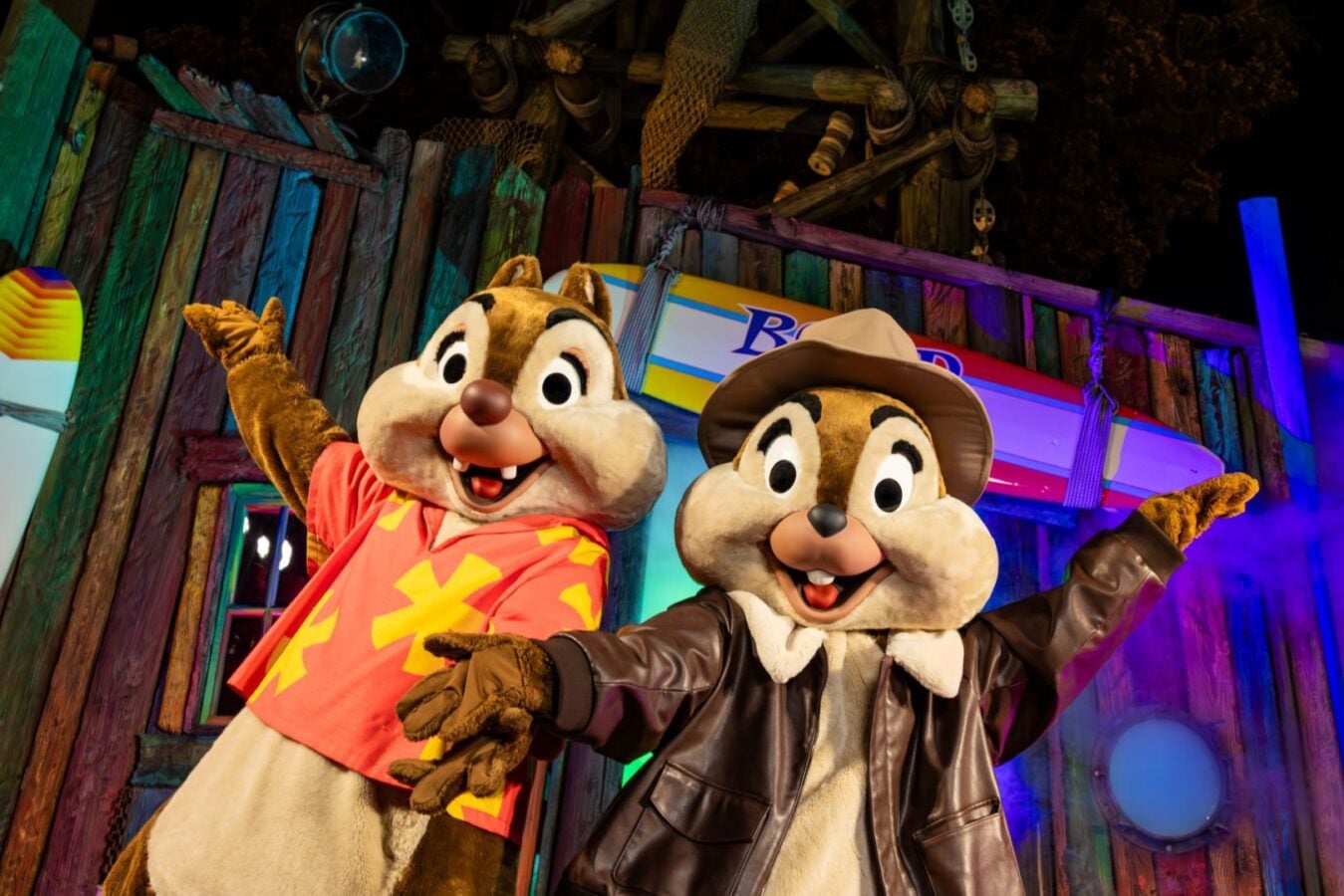 Two Chip n Dale mascots pose with arms raised in a bright tropical themed setting featuring a colorful wooden wall and a surfboard in the background