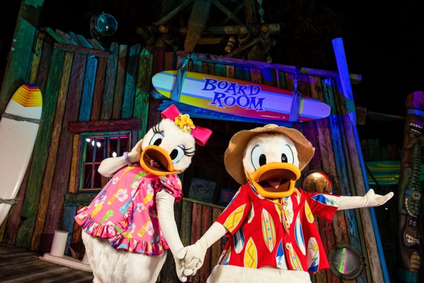 Two costumed ducks in bright tropical clothing hold hands on a colorful wooden stage with a Board Room sign above them suggesting a beachy playful theme