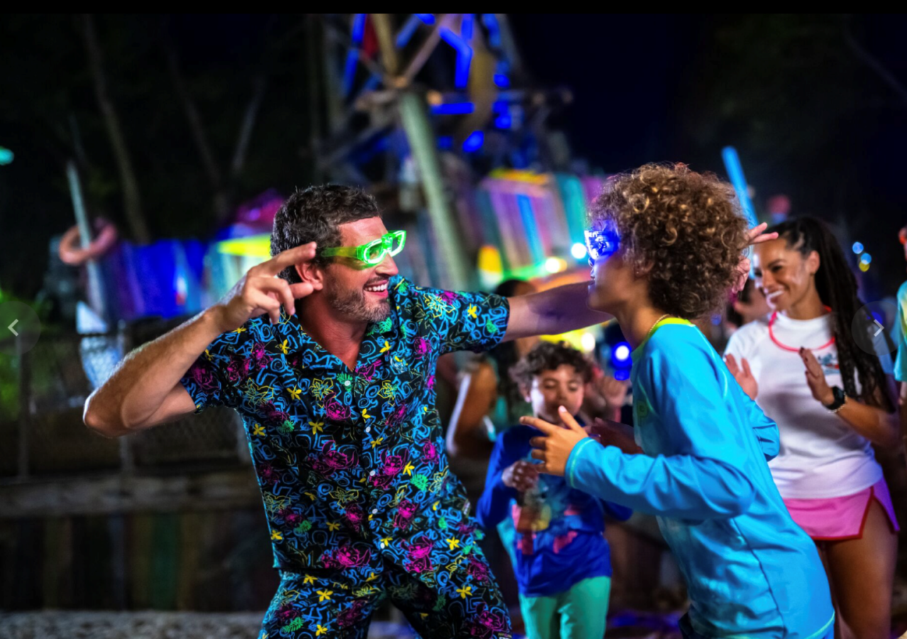Man and boy dancing at a neon lit party wearing glowing green sunglasses and a colorful hawaiian shirt
