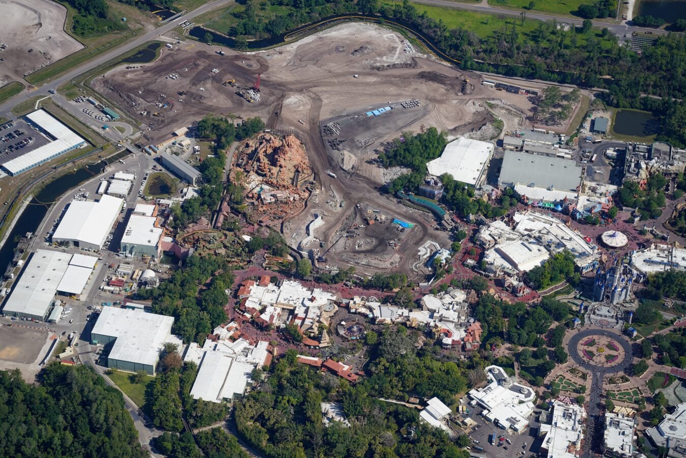 Aerial view of a large construction site in an urban area with dirt piles heavy machinery and cleared ground surrounded by roads and buildings A circular plaza with greenery is visible toward the bottom right