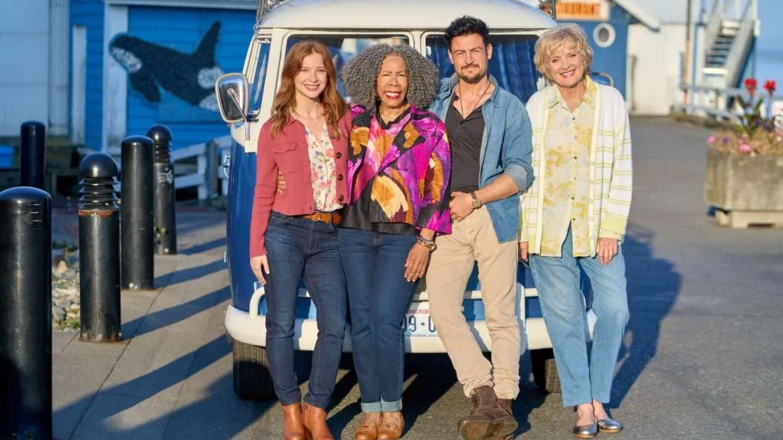 Four adults smile together while leaning against a vintage white van on a sunny seaside boardwalk