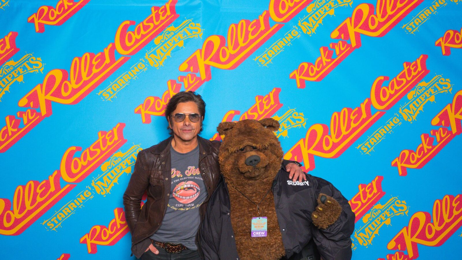 JohnStamosandTheMuppets Fantasy Land News Man in sunglasses and a brown jacket posing with a brown bear mascot against a blue backdrop with Rockn Roller and The Muppets logos used for a photo op