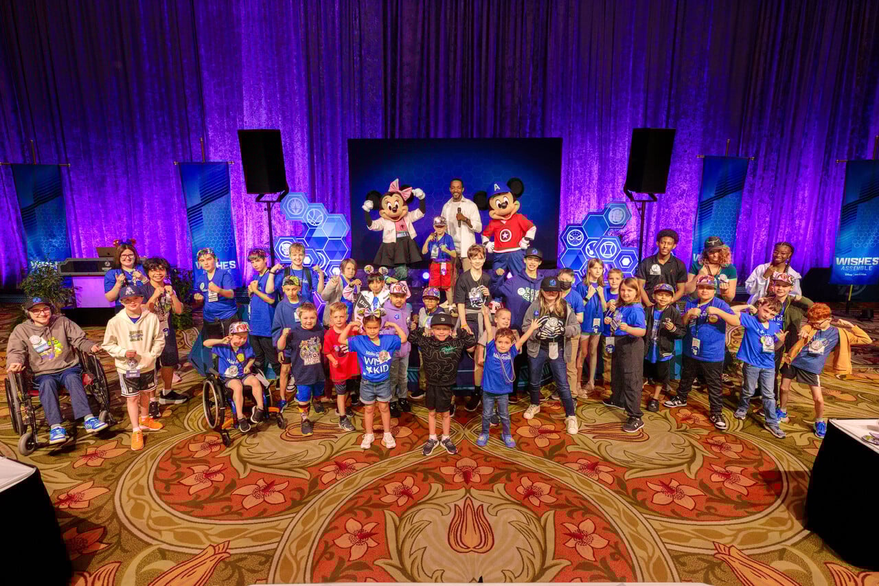 Group of children and adults posing for a photo on a stage with costumed characters under purple lighting in a large event hall