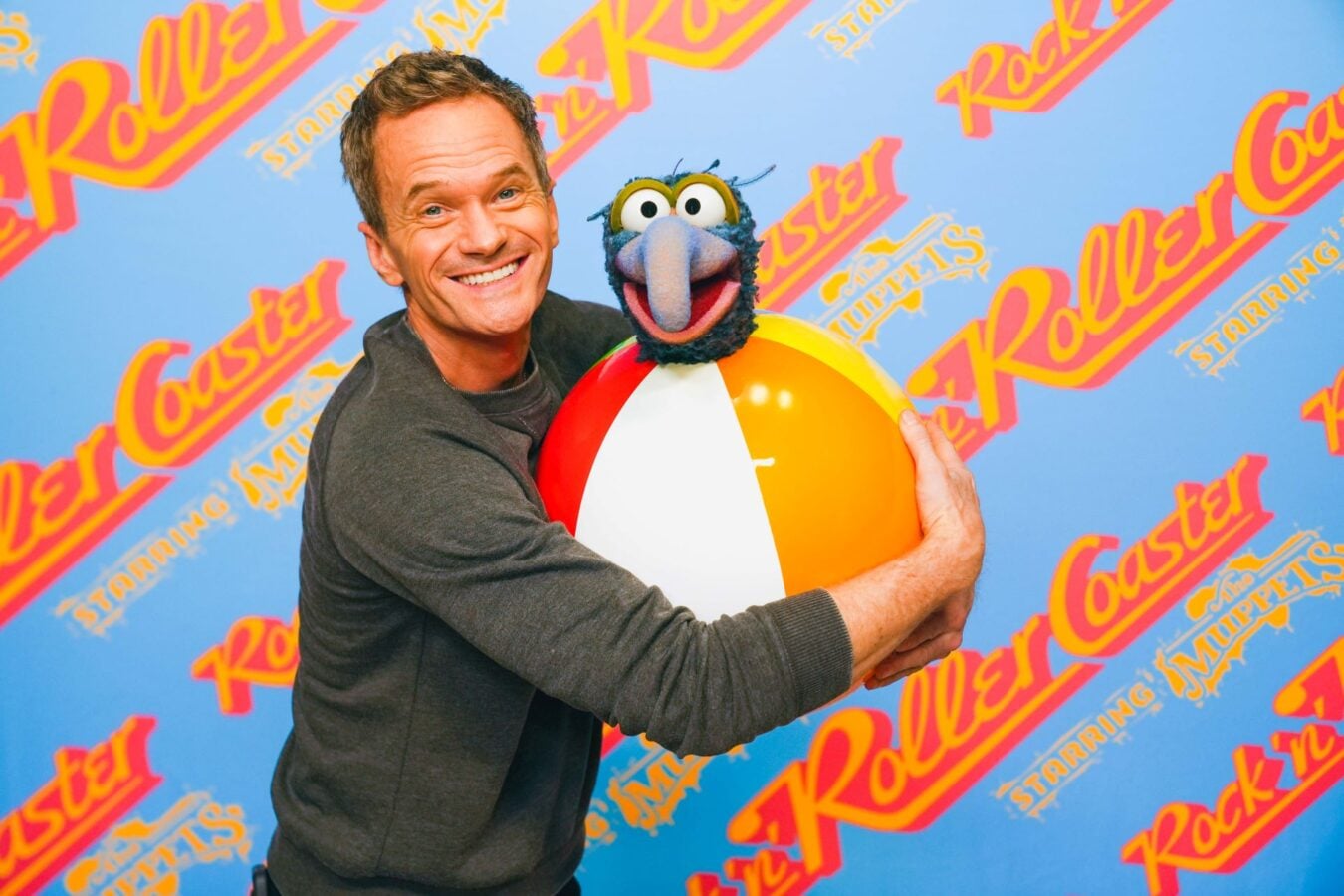 NeilPatrickHarriswithGonzoandTheMuppets Fantasy Land News Man smiles while hugging a large beach ball with a puppet head on top against a blue poster wall with Muppets branding