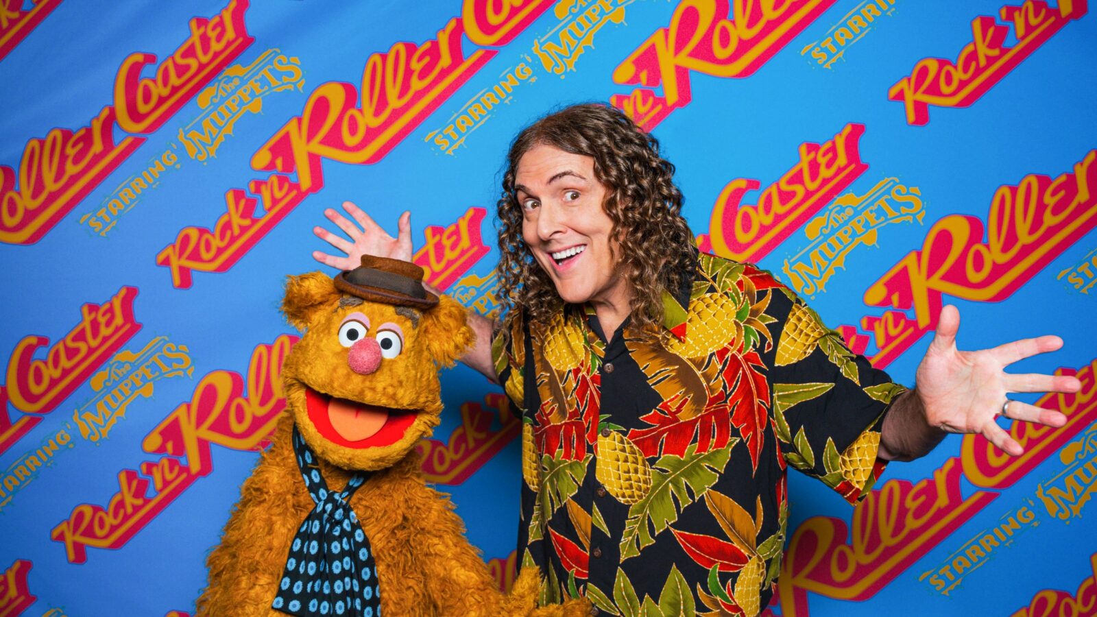 RnRFozzieAlYankovicscaled Fantasy Land News Man with long curly hair in a tropical shirt poses with a furry orange Muppet wearing a hat against a bright blue backdrop filled with Roller Rockin and The Muppets logos