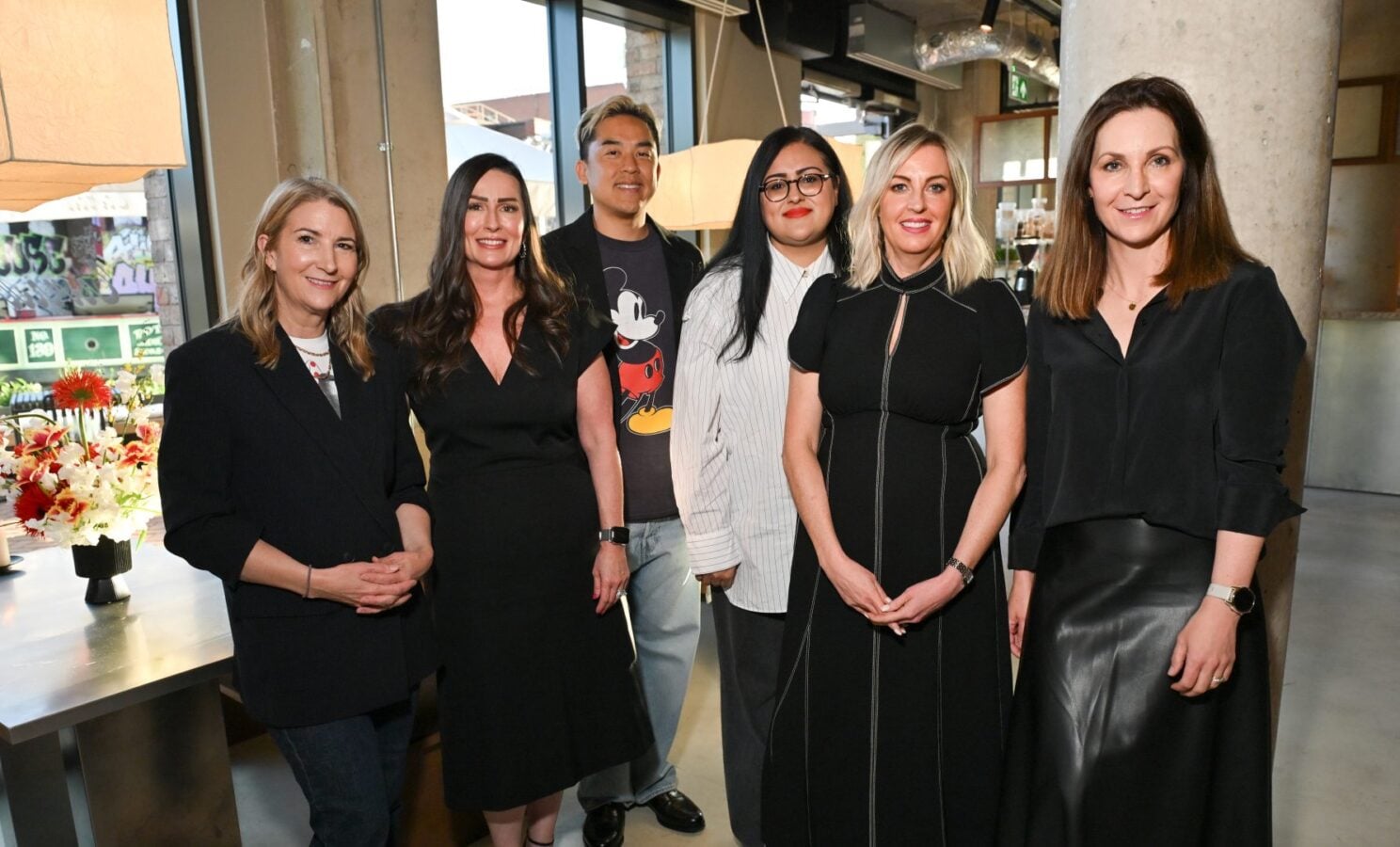 Six people stand together indoors smiling for the camera in a modern industrial style space wearing business casual attire suggesting a group photo at a work event