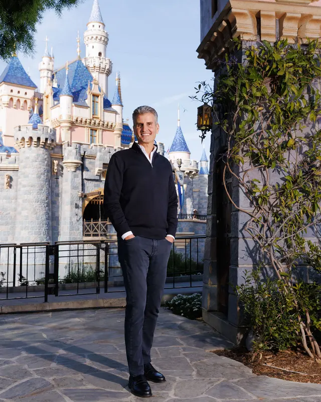 T1002026WebJoshDAmaro Fantasy Land News Man in a black sweater and dark jeans stands with hands in pockets on a stone walkway with a fairytale castle featuring blue roofed towers in the background on a sunny day