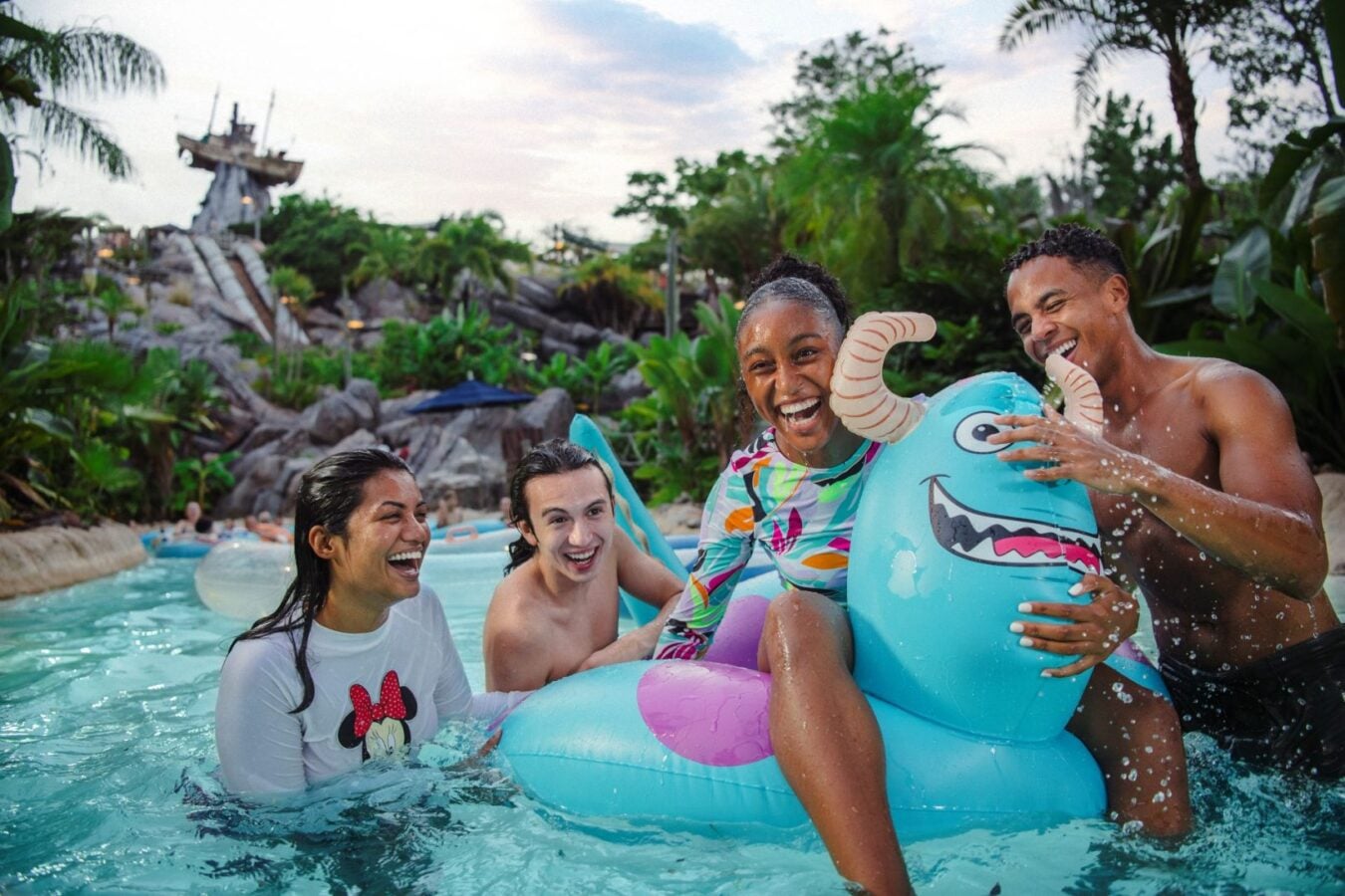 Four friends laughing and riding a blue inflatable dinosaur in a tropical pool setting