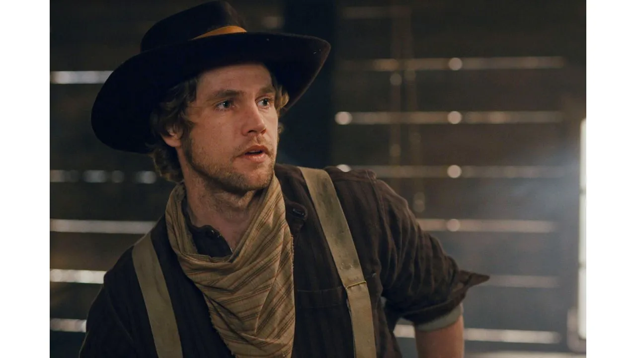 Man in a wide brimmed hat scarf and suspenders in a dim wooden western setting looking off to the side