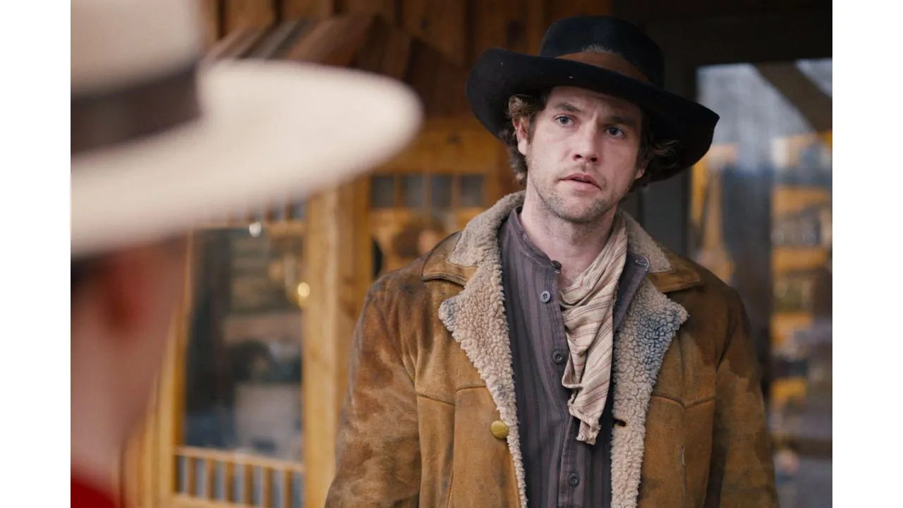 Man in a brown shearling coat and black cowboy hat looks off to the side outdoors with a wooden building in the background