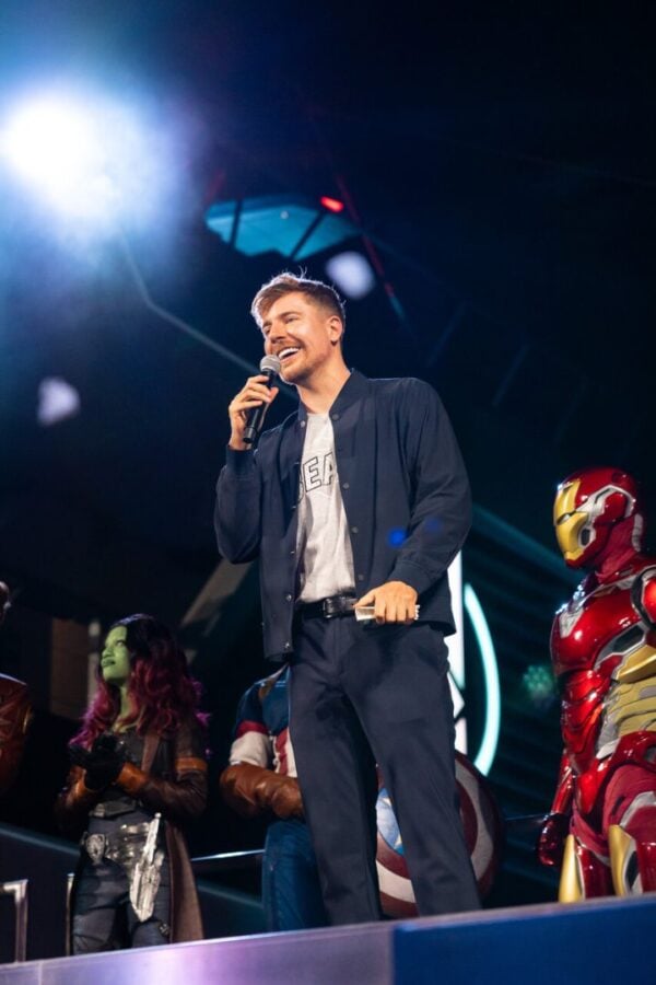Male performer in a navy jacket sings into a handheld microphone on a stage with bright lights actors in costumes behind him