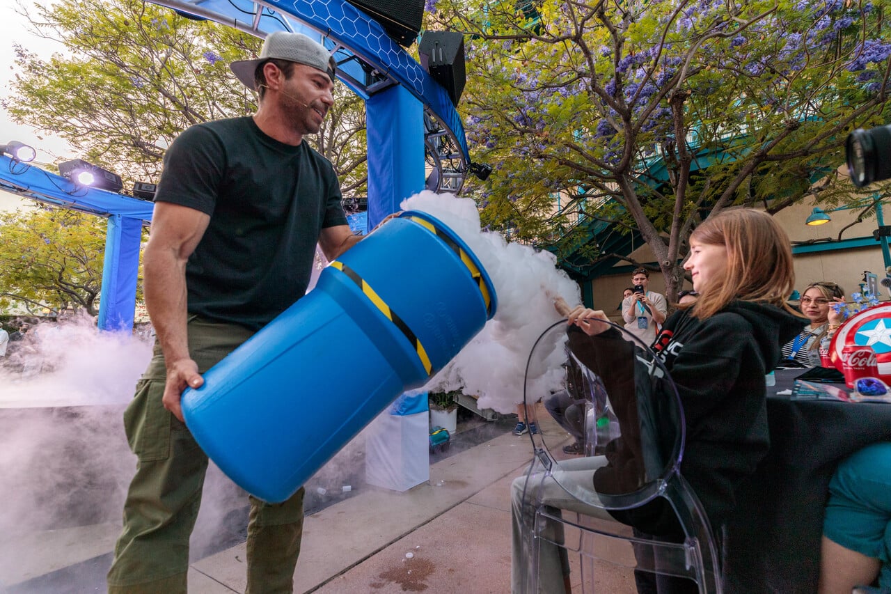 Man in a black t shirt uses a blue fog machine to emit vapor toward a seated girl at an outdoor event