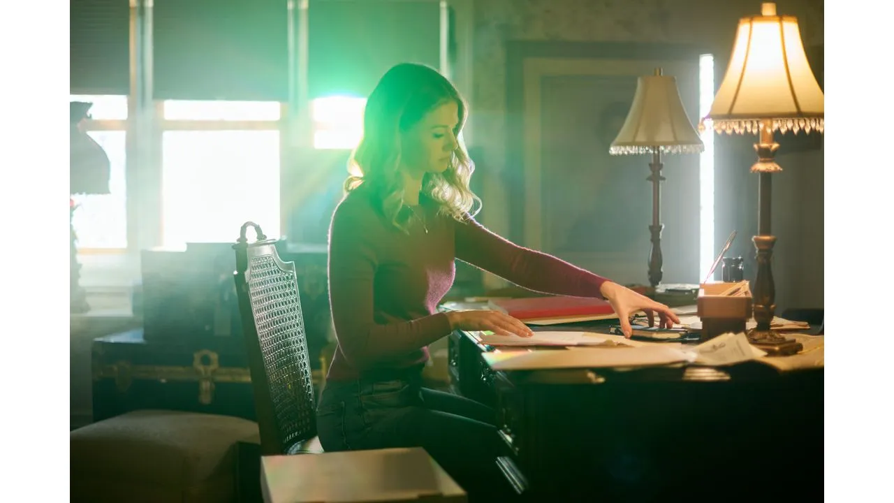 Woman sits at a vintage desk sorting papers by warm lamp light in a sunlit room