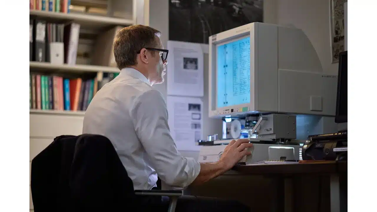 Man in a white shirt and glasses sits at a vintage computer terminal typing at a large monitor in a book filled office