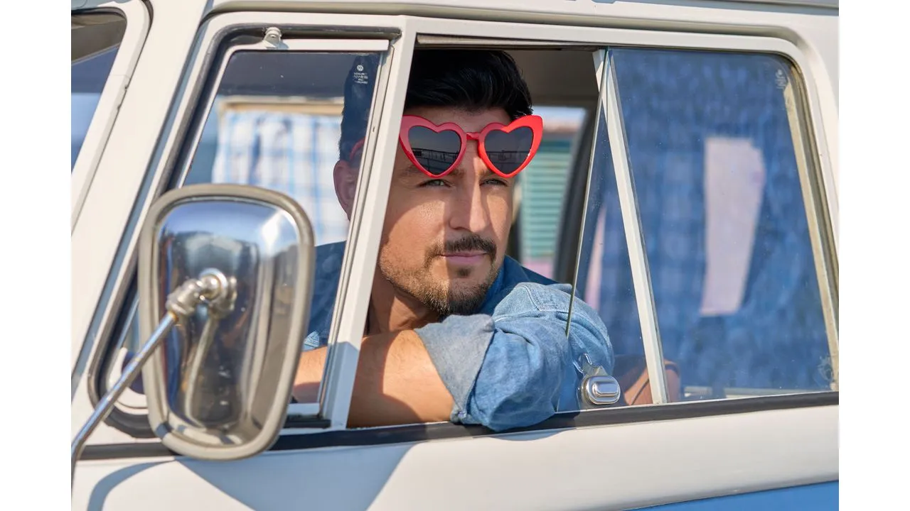 Man with a trimmed beard sits in the drivers seat of a vintage white car wearing red heart shaped sunglasses and a blue denim shirt gazing out the window'I'll Be Seeing You' Premieres Tonight on Hallmark Channel