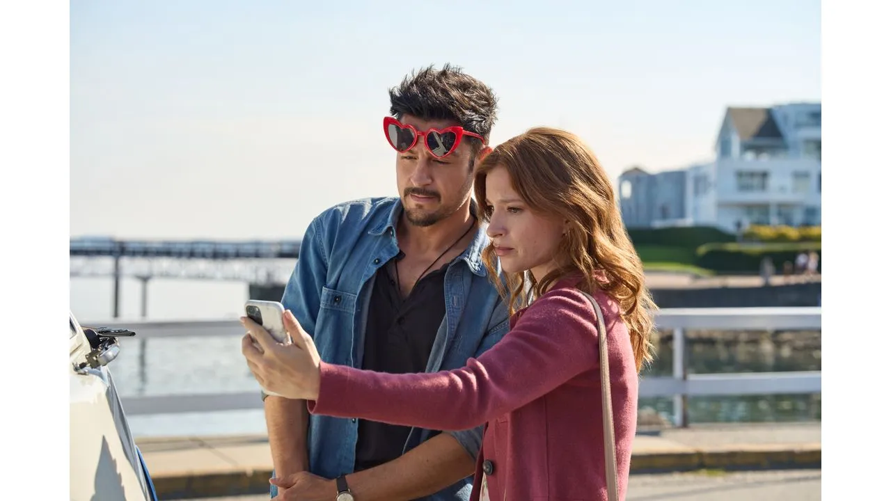 Couple takes a selfie by the water with a phone woman in pink coat and man in denim shirt wearing red heart sunglasses nearby a parked car by a pier blue sky behind them
