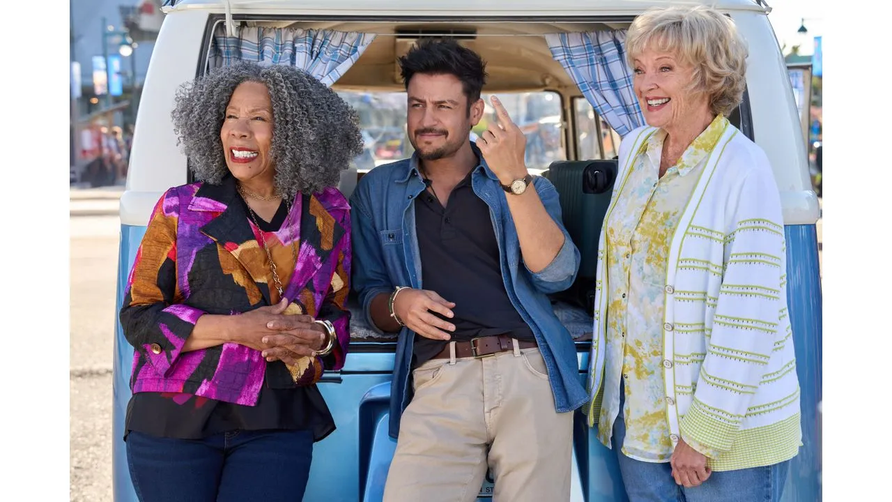 Three smiling friends pose by a retro blue van with checkered curtains in the doorway