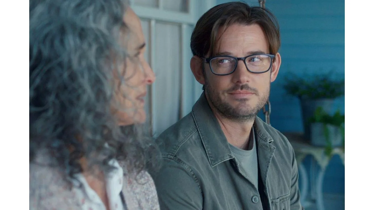 Two men talking on a porch the man on the right wears glasses and a green jacket looking at his gray haired companion who is blurred in the foreground