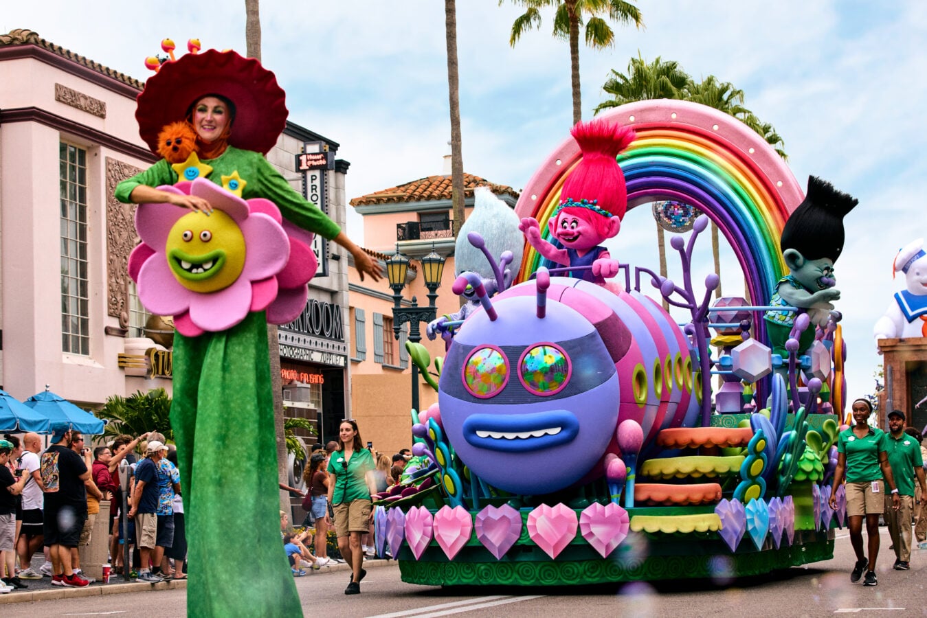 Colorful parade float with a pink haired Troll mascot on a rainbow colored vehicle and oversized pink flower crowds line the street to watch