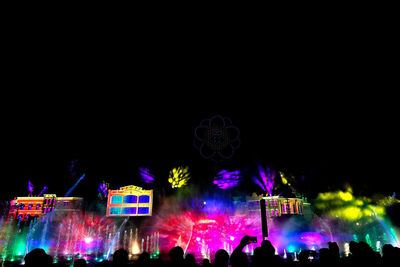 Nighttime fountain show with multicolored lights silhouetted crowd in the foreground and a glowing circular light pattern in the sky