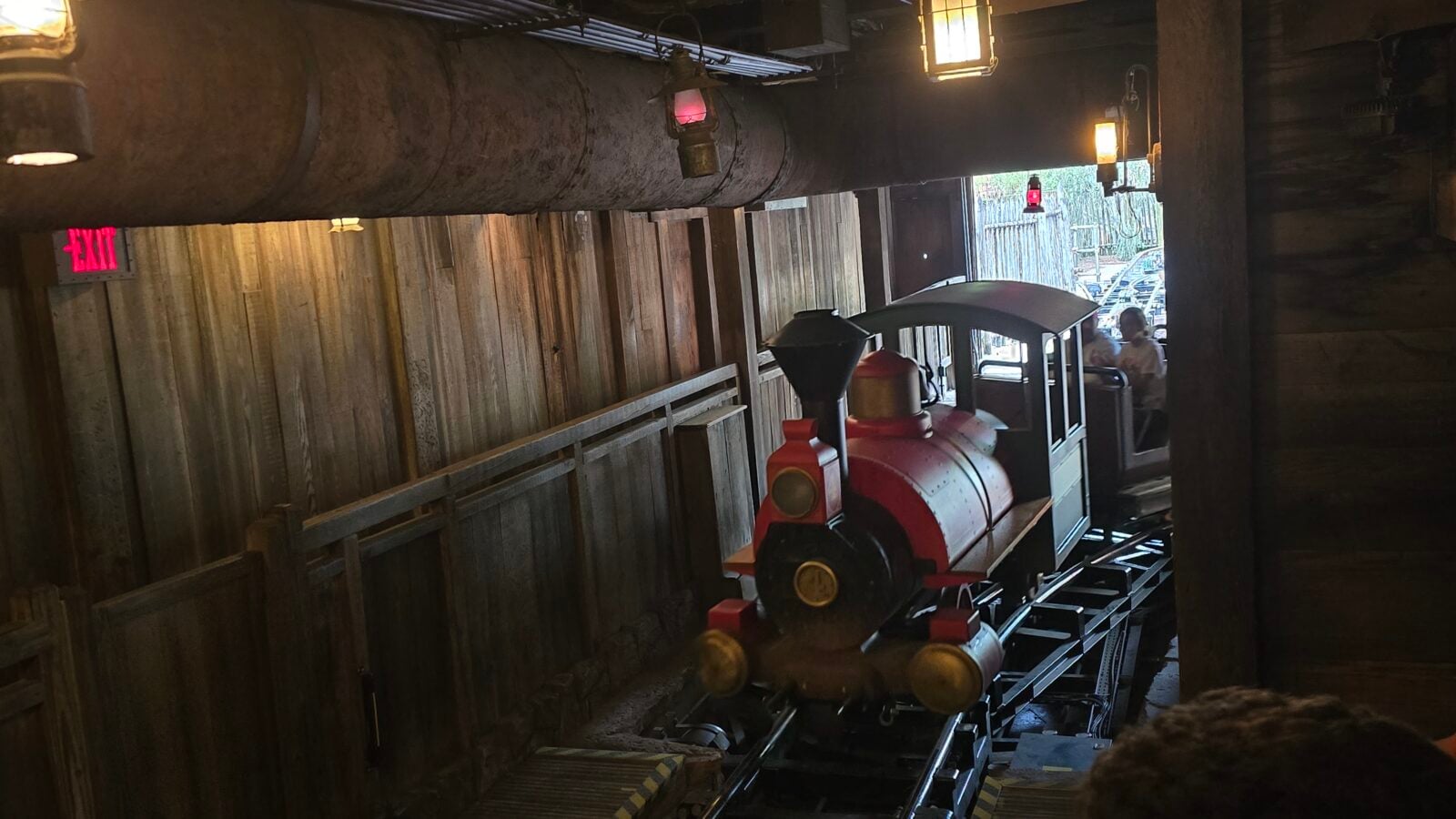 Interior of a rustic mine train ride red locomotive on tracks beside wooden walls with hanging lanterns and an illuminated exit sign in the background