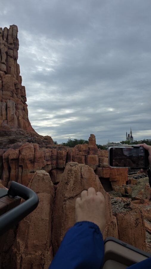 Person in a blue jacket holding a smartphone to photograph tall red rock formations under a cloudy sky with a distant castle on the horizon