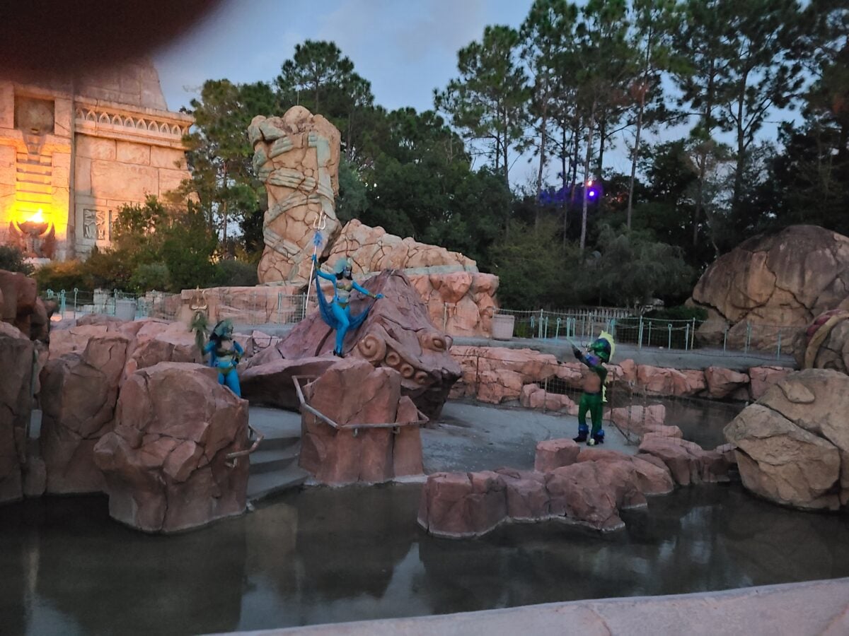 Outdoor stage with a large artificial rock sculpture and performers in bright blue outfits near a water feature at dusk