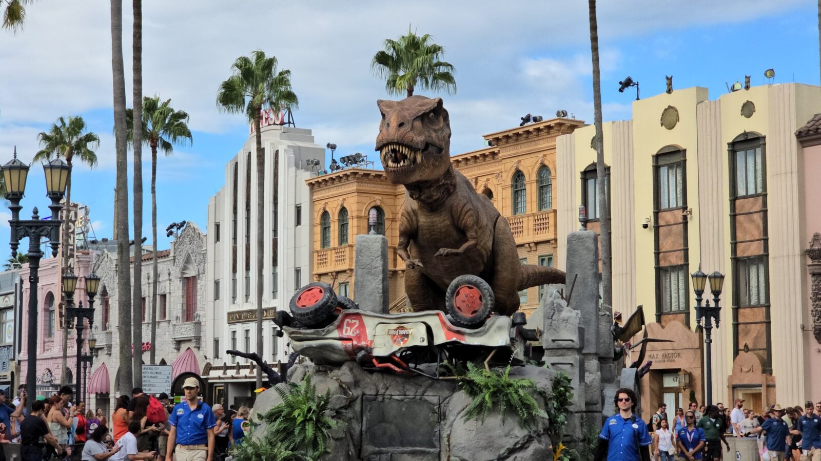 Life size dinosaur statue atop a crashed race car in a theme park street with palm trees and pastel buildings in the background and spectators nearby