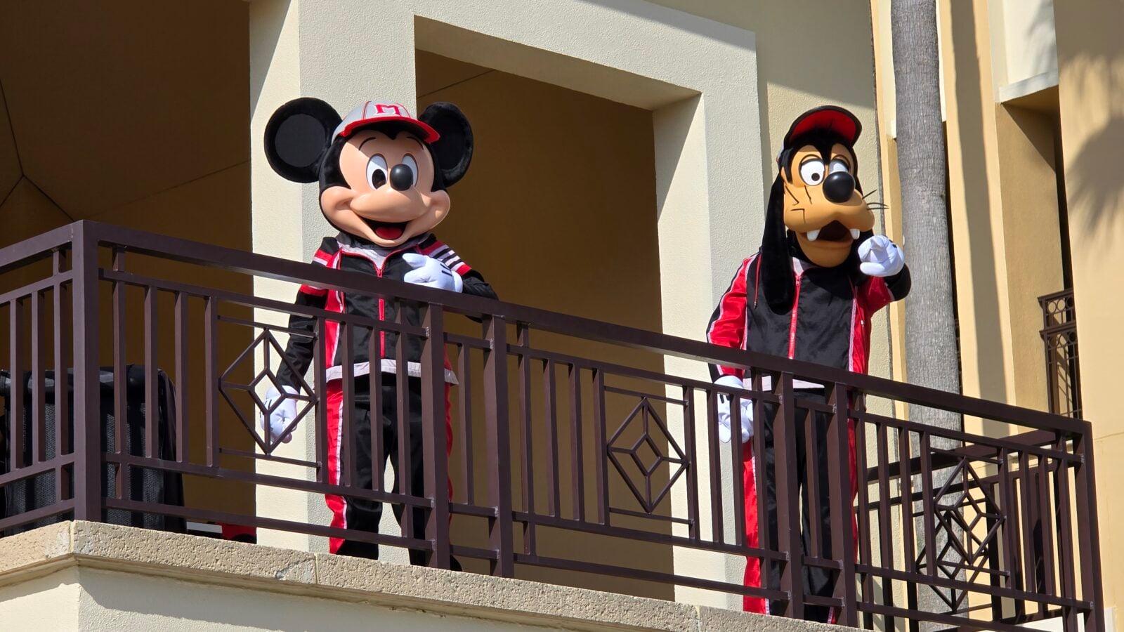 Mickey Mouse and Goofy mascots in red black outfits stand on a balcony railing of a sunny beige building