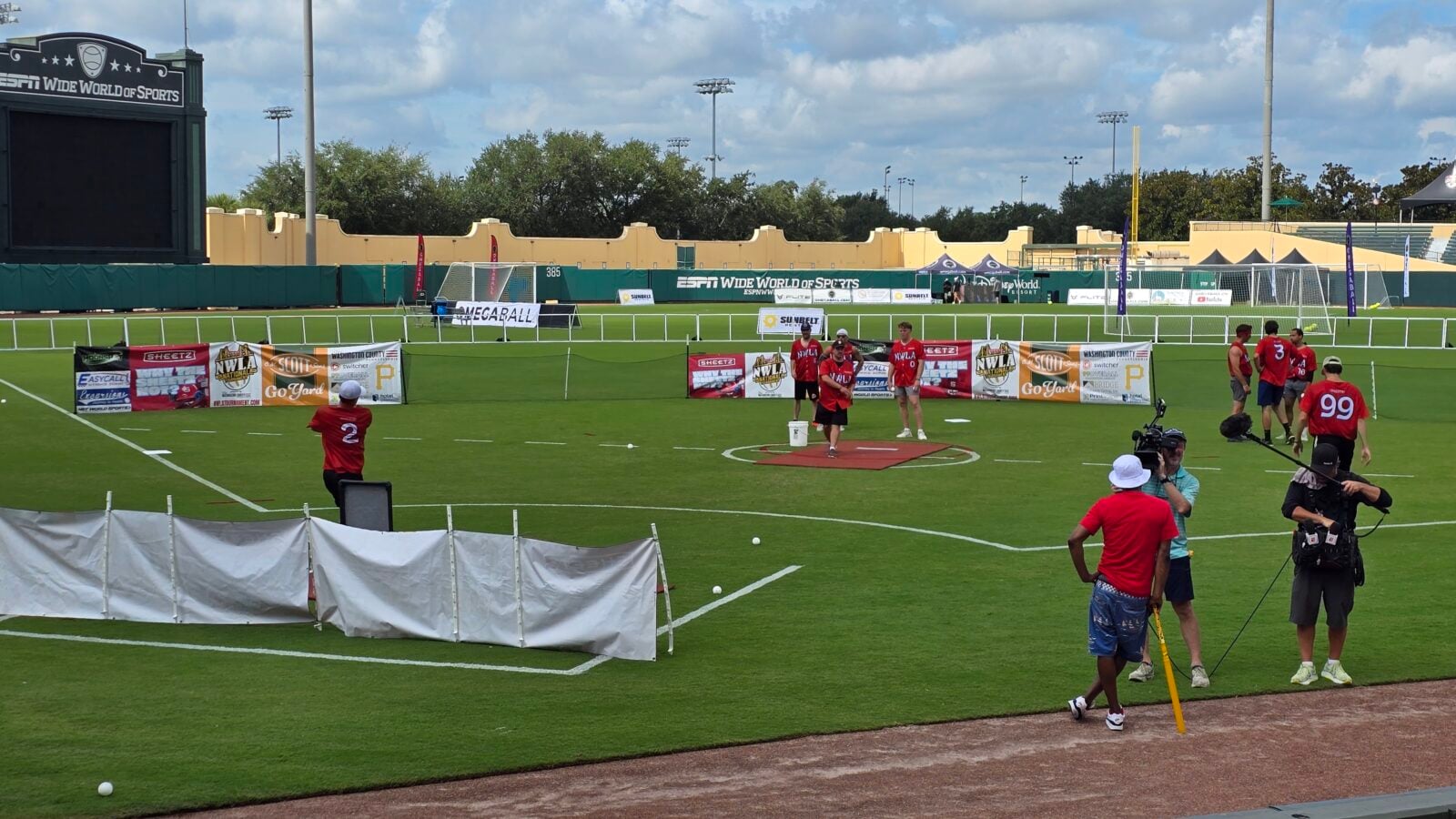 Group of athletes in red jerseys standing on a green sports field near advertising banners and a barrier wall with a camera crew in the foreground