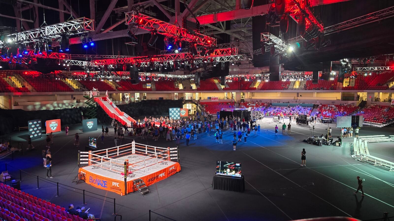Boxing ring in a large arena with red ropes surrounded by crew and scattered spectators under red and blue overhead lights