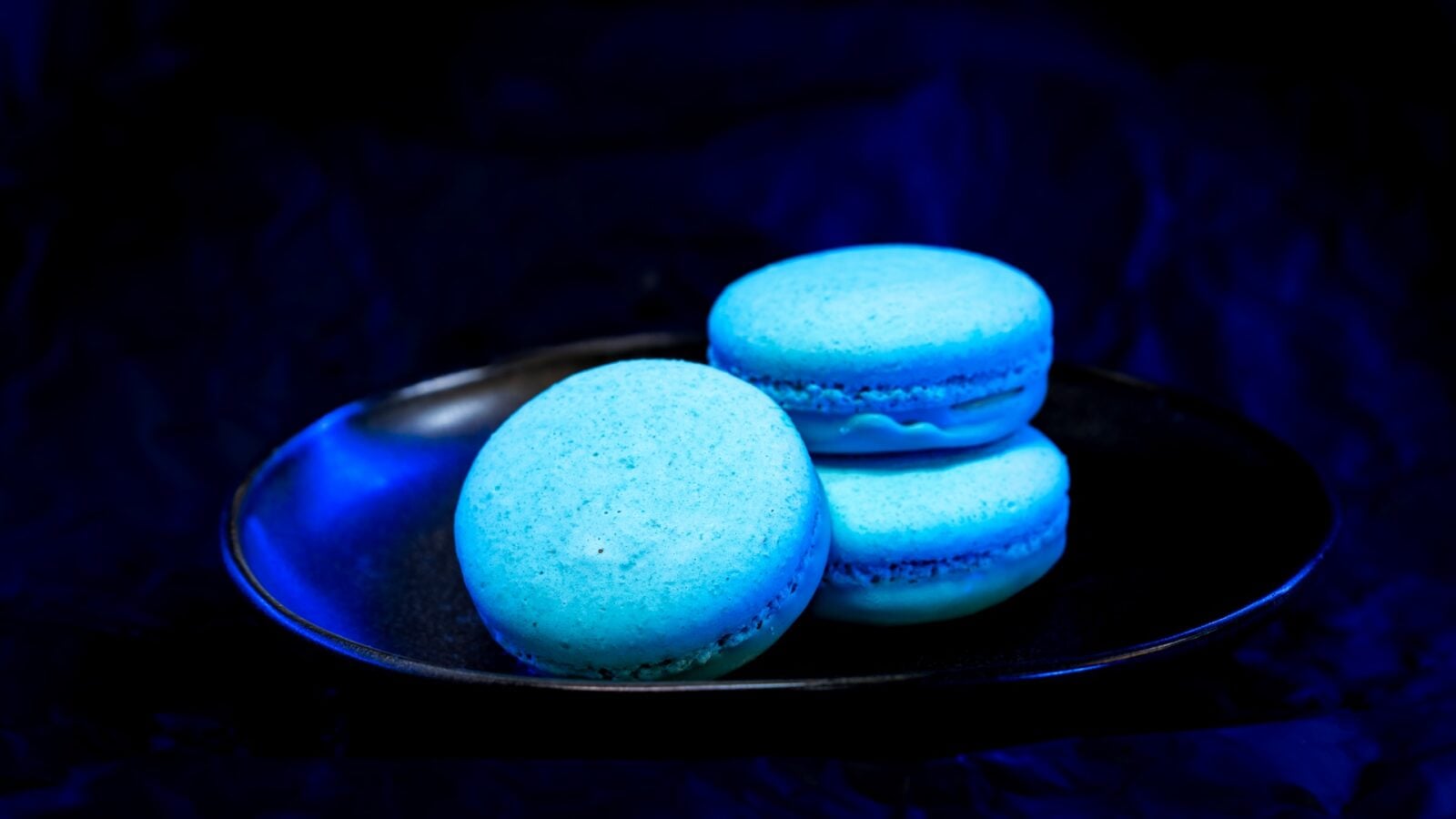 Blue macarons stacked on a dark plate with blue lighting in a moody setting