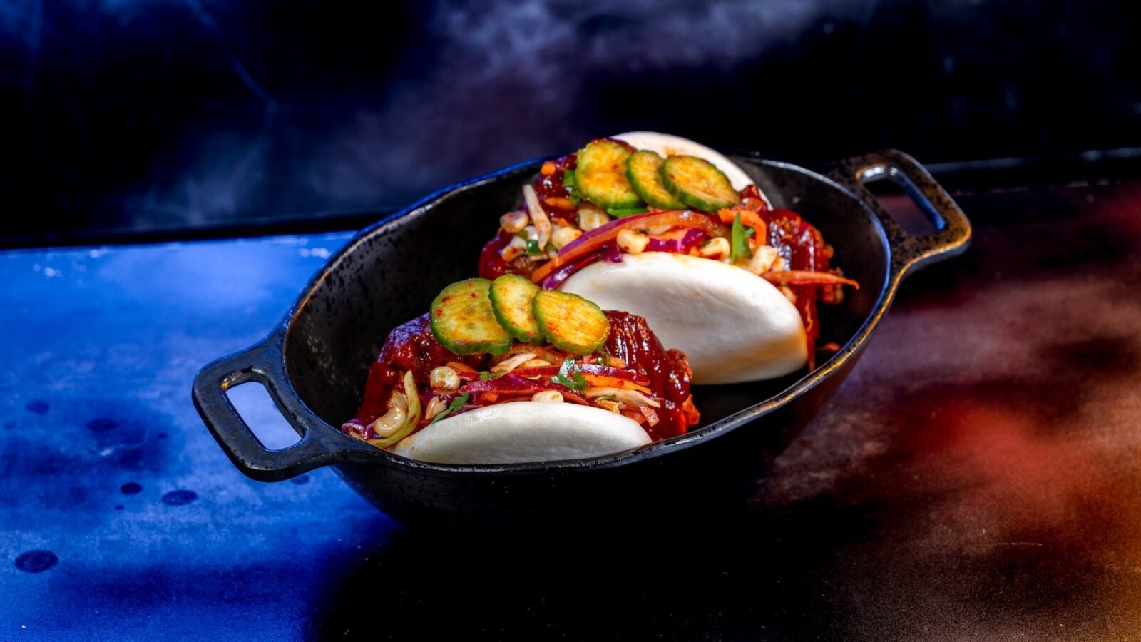 Two soft tacos in a black skillet filled with red sauce corn onions peppers and cucumber slices on top