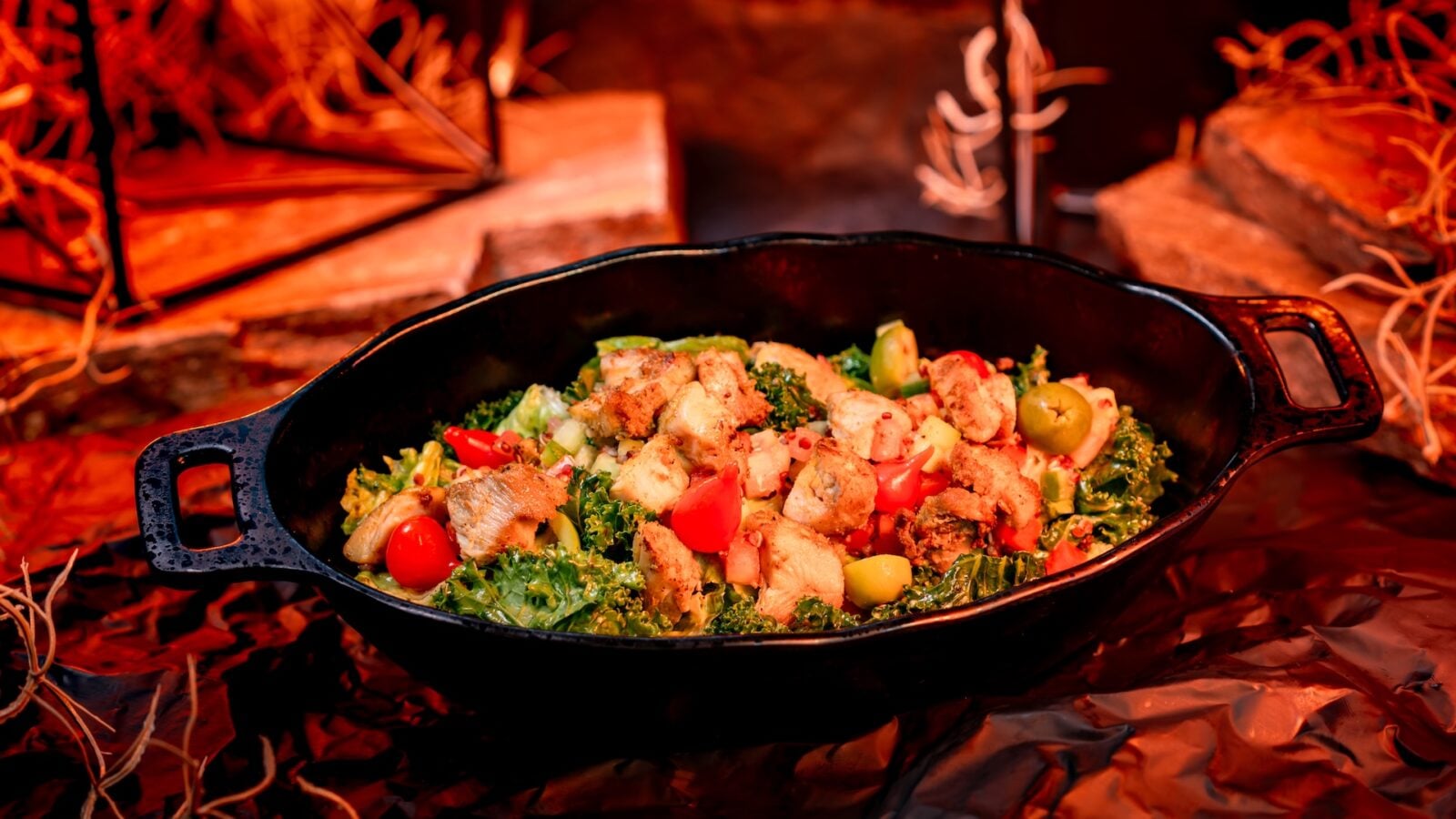 Cast iron skillet of chicken salad with greens peppers olives and cherry tomatoes cooking over a campfire