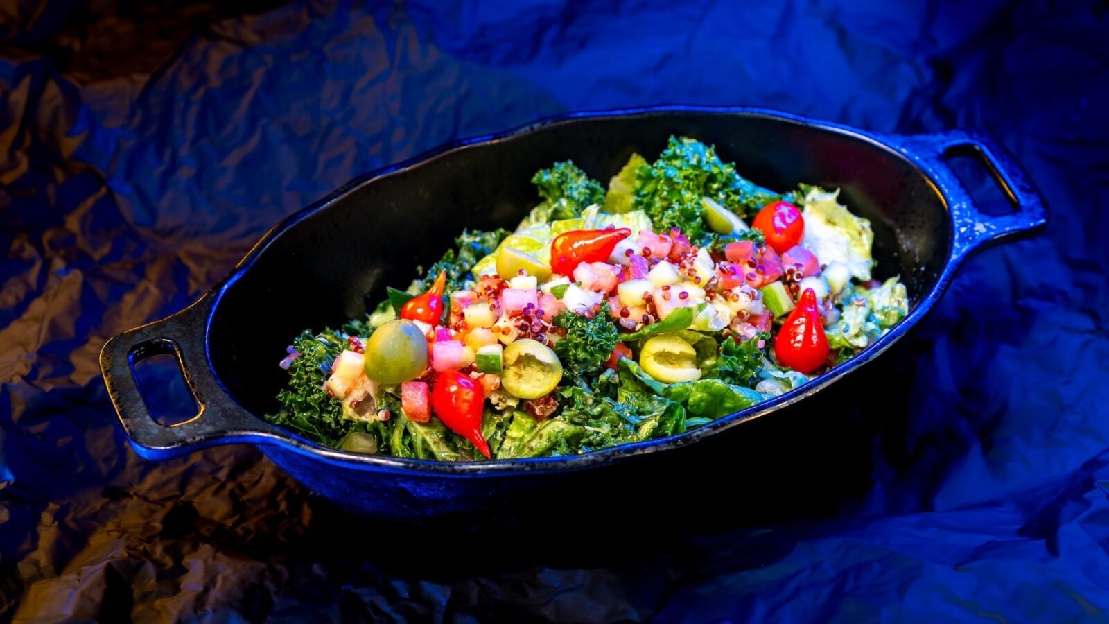Colorful chopped salad with kale cucumbers olives peppers and tomatoes in a black cast iron skillet on a dark blue background